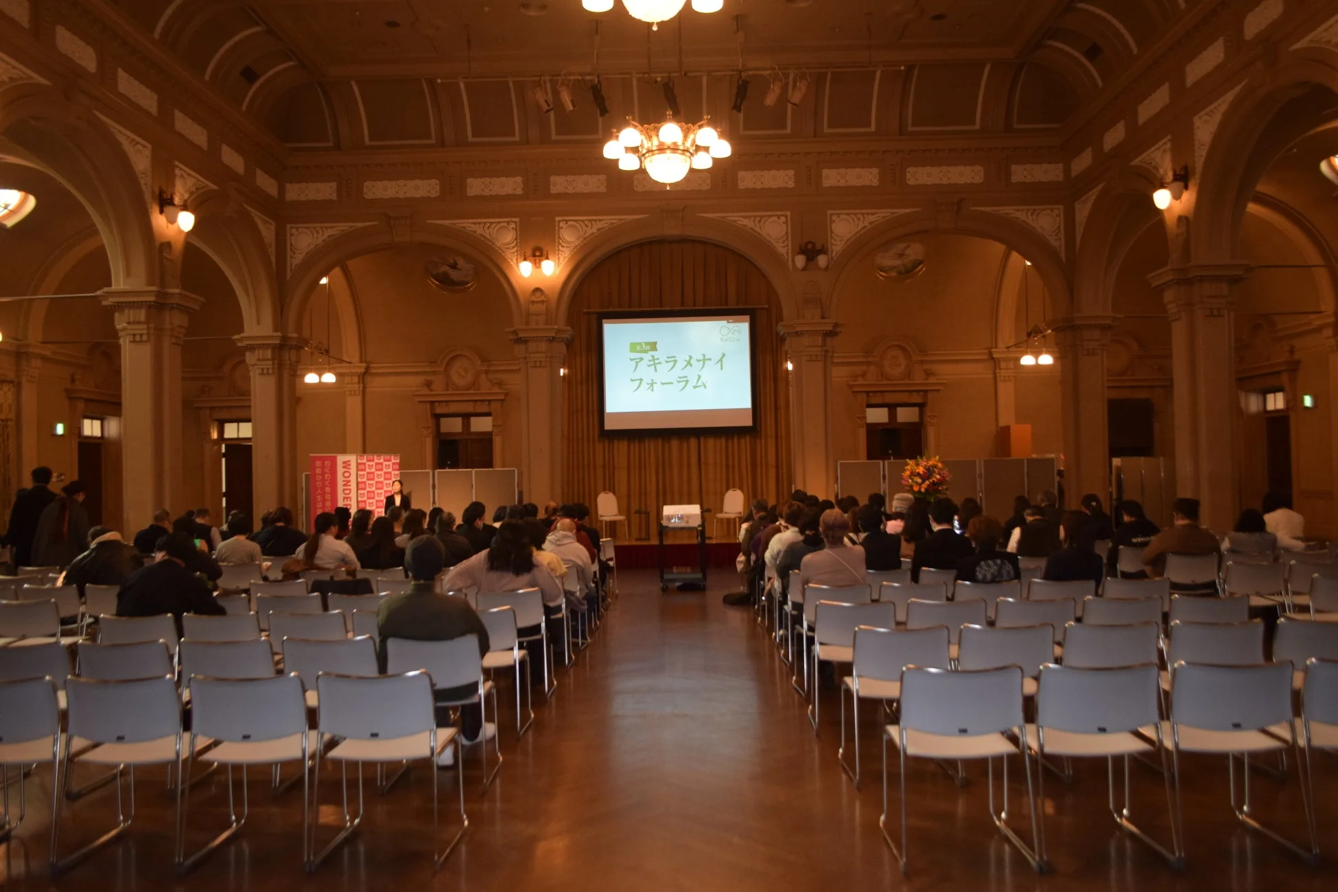 豪華な装飾が施された広間で行われているフォーラムの様子です。多くの参加者が椅子に座り、正面のスクリーンに映し出された「アキラメナイ フォーラム」という文字を見ています。歴史的な建造物で開催されているイベントのようです。