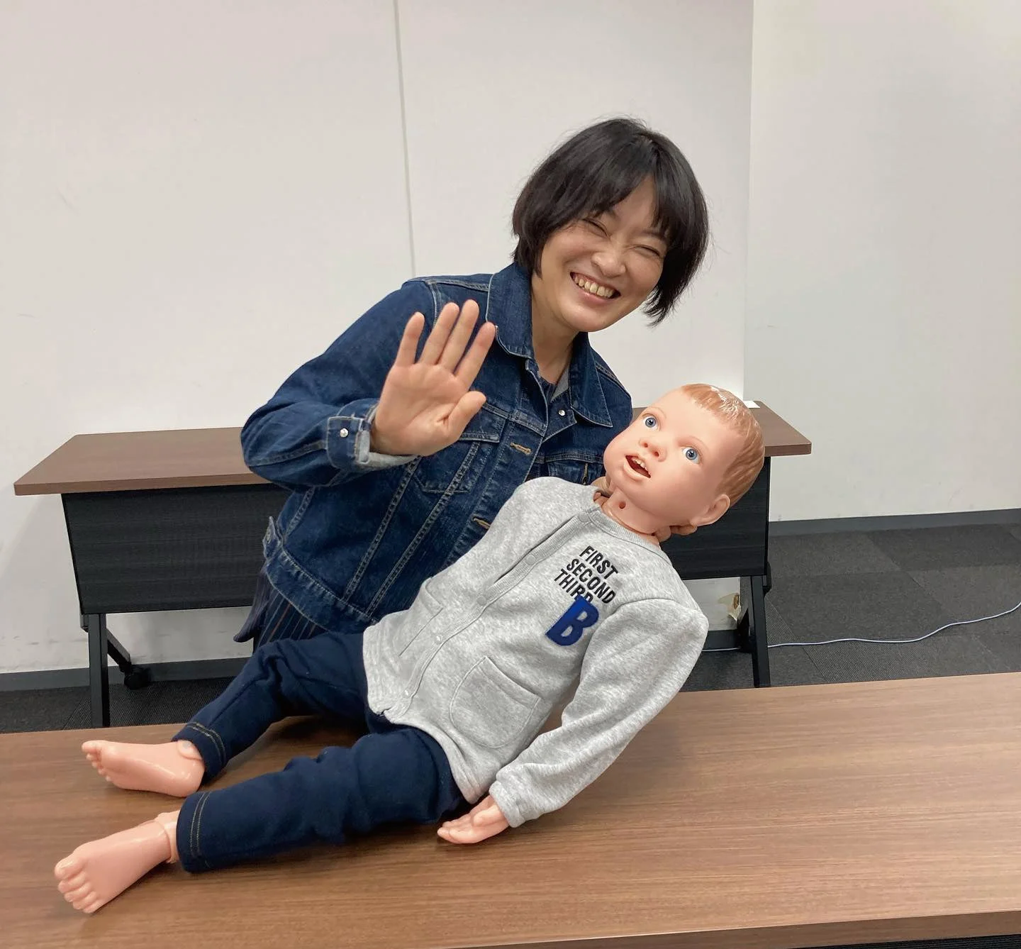 笑顔の女性が机に横たわる赤ちゃんの人形と一緒に写っている