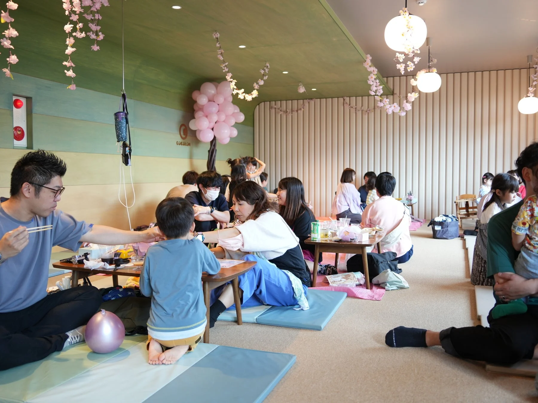 桜とバルーンで飾られた明るい室内で、多くの家族連れが低いテーブルを囲んで食事やくつろぎの時間を過ごしています。