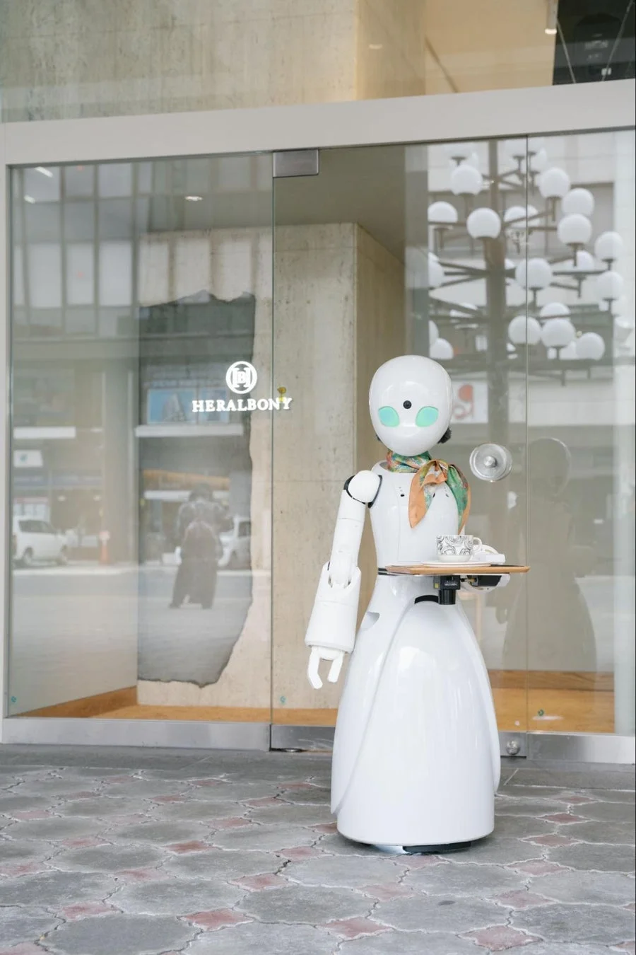 白いサービスロボットが、ガラス張りの建物の入り口前でトレイにカップを載せて立っています。