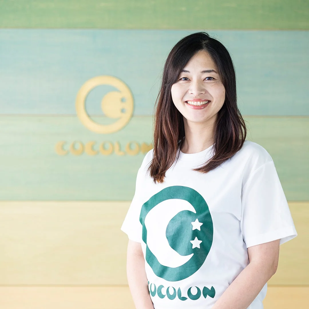 COCOLONのロゴ入りTシャツを着た女性が笑顔で立っています。