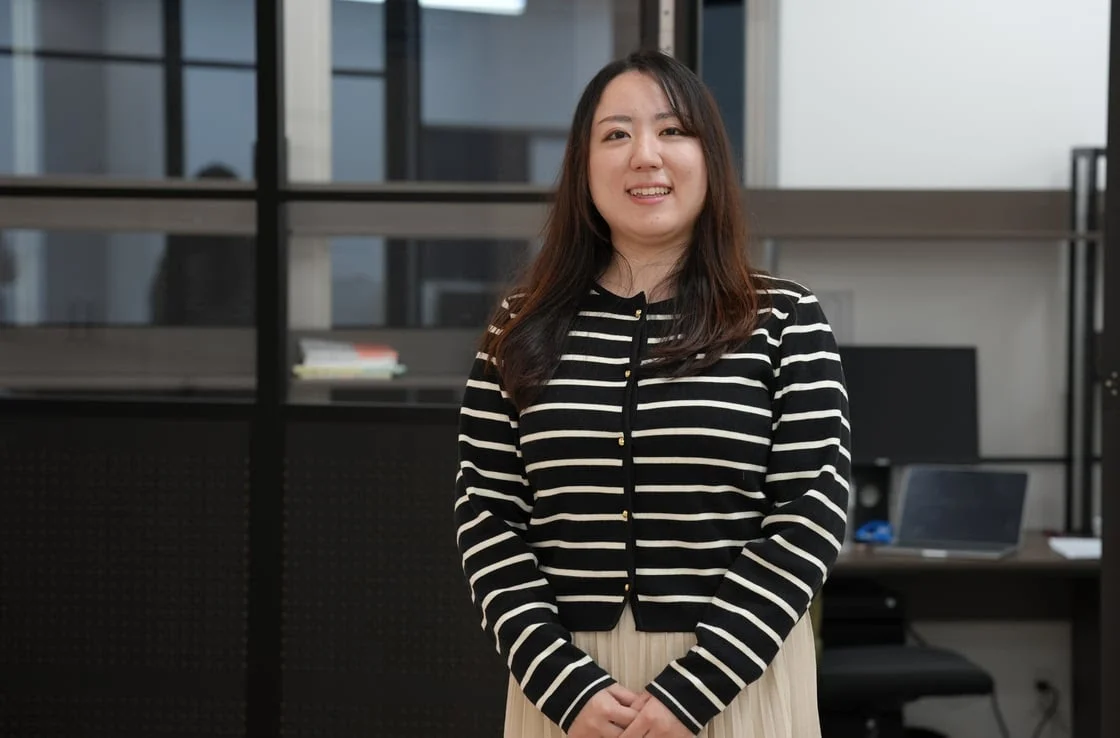 オフィスのような室内で、黒と白のストライプのカーディガンを着た笑顔の若い女性が立っています。