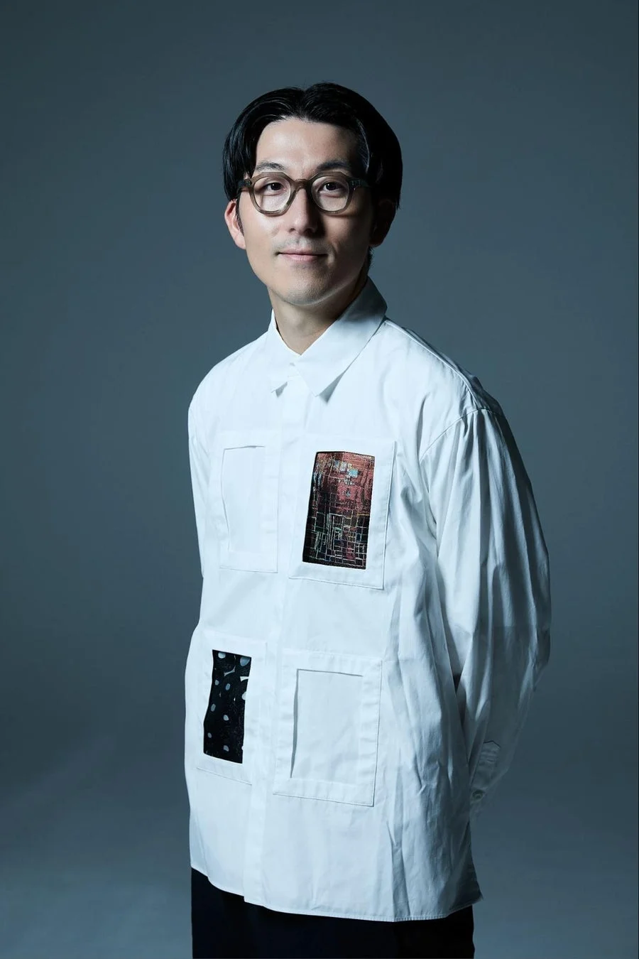 眼鏡をかけたアジア人男性が、ユニークなパッチワークデザインの白いシャツを着て、カメラに向かって穏やかに微笑んでいるポートレート写真です。
