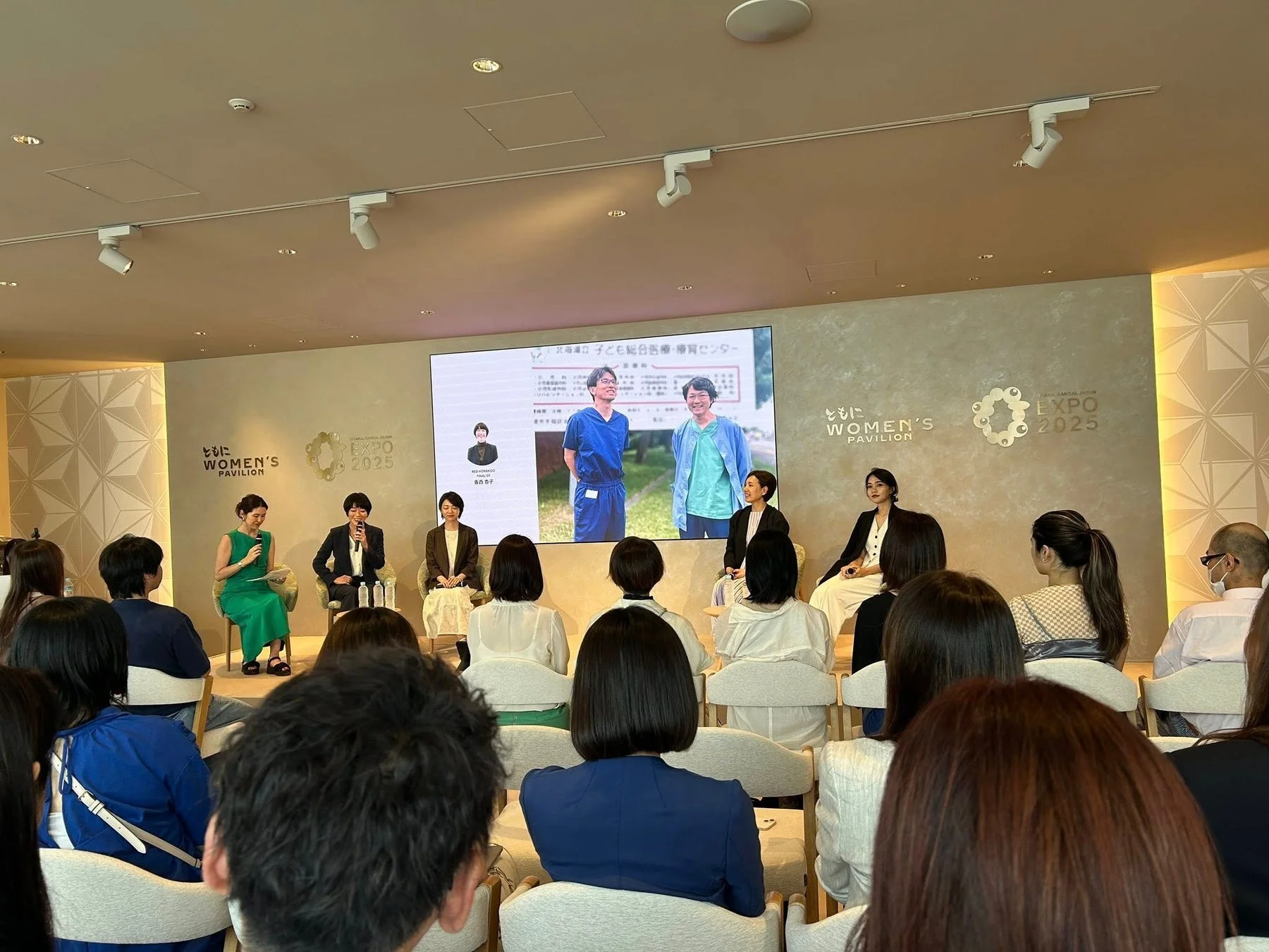 「WOMEN'S PAVILION」と「EXPO 2025」関連イベントでのパネルディスカッション風景。女性登壇者が聴衆の前で話し、スクリーンには「こども総合診療・療育センター」の情報が表示されている。