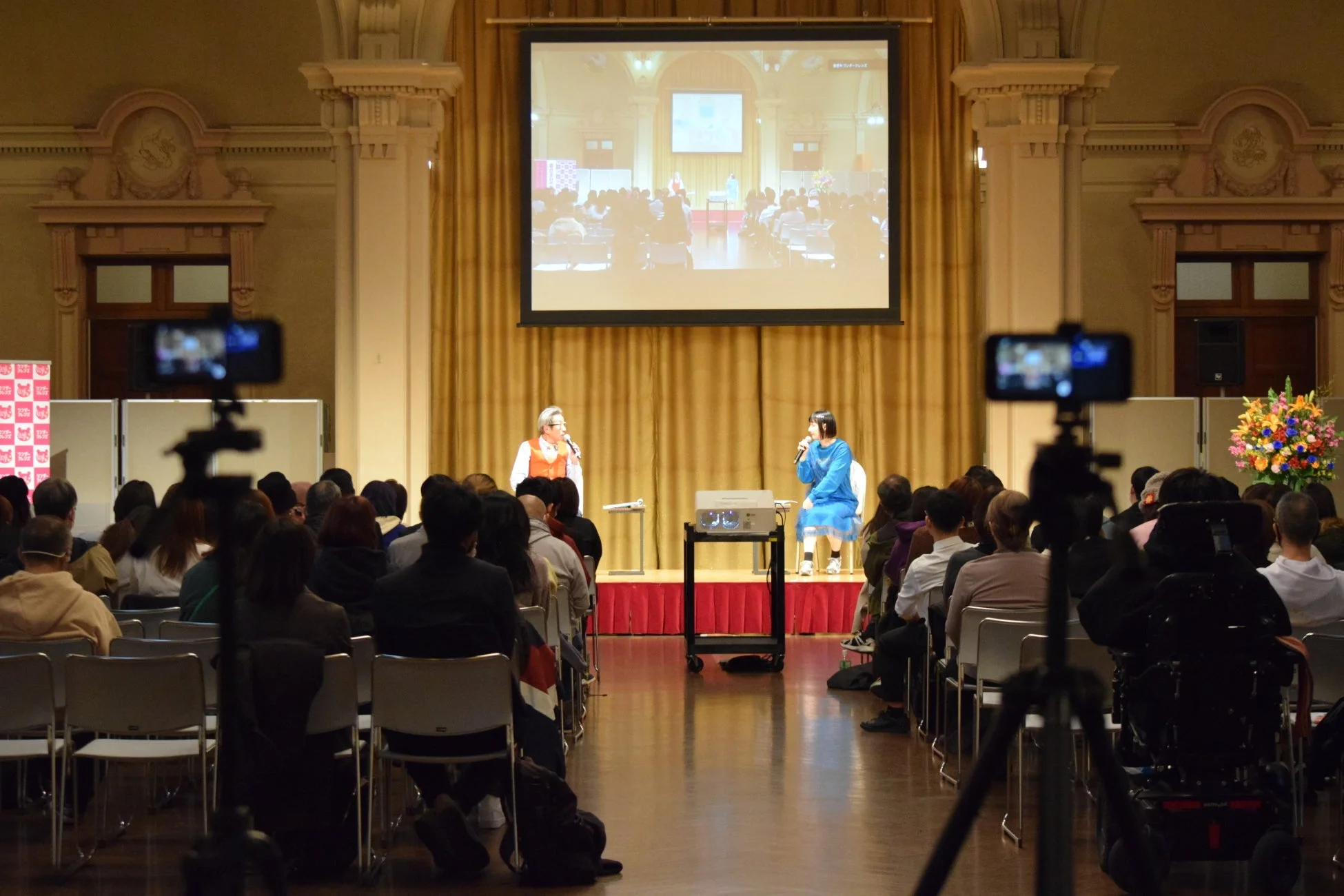 室内で行われている講演会またはセミナーの様子です。ステージ上の登壇者2名がマイクで話しており、後方の大画面にはその様子が映し出されています。会場には多くの聴衆が着席し、複数のカメラで撮影が行われています。