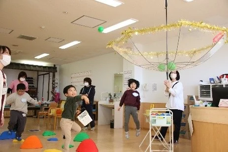 室内で子供たちが大人たちと楽しそうに遊んでいる様子。一人の男の子がボールを投げ、逆さにした傘を的にしている。他の子供も飛び跳ねたり、色とりどりの踏み台の上を歩いたりしており、活気あふれる遊びの光景です。