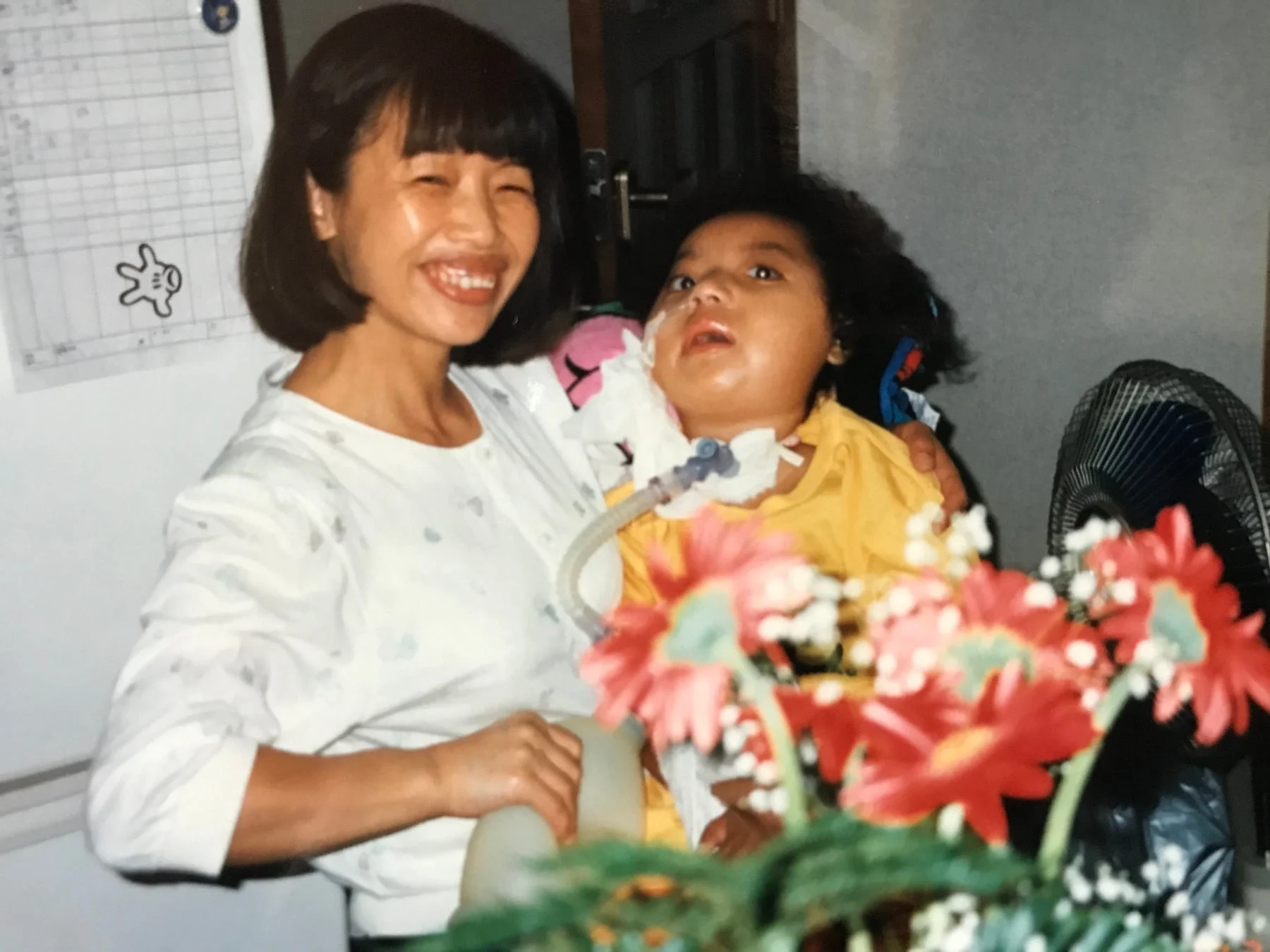 笑顔の女性が医療チューブを装着した子供を抱きかかえている室内写真