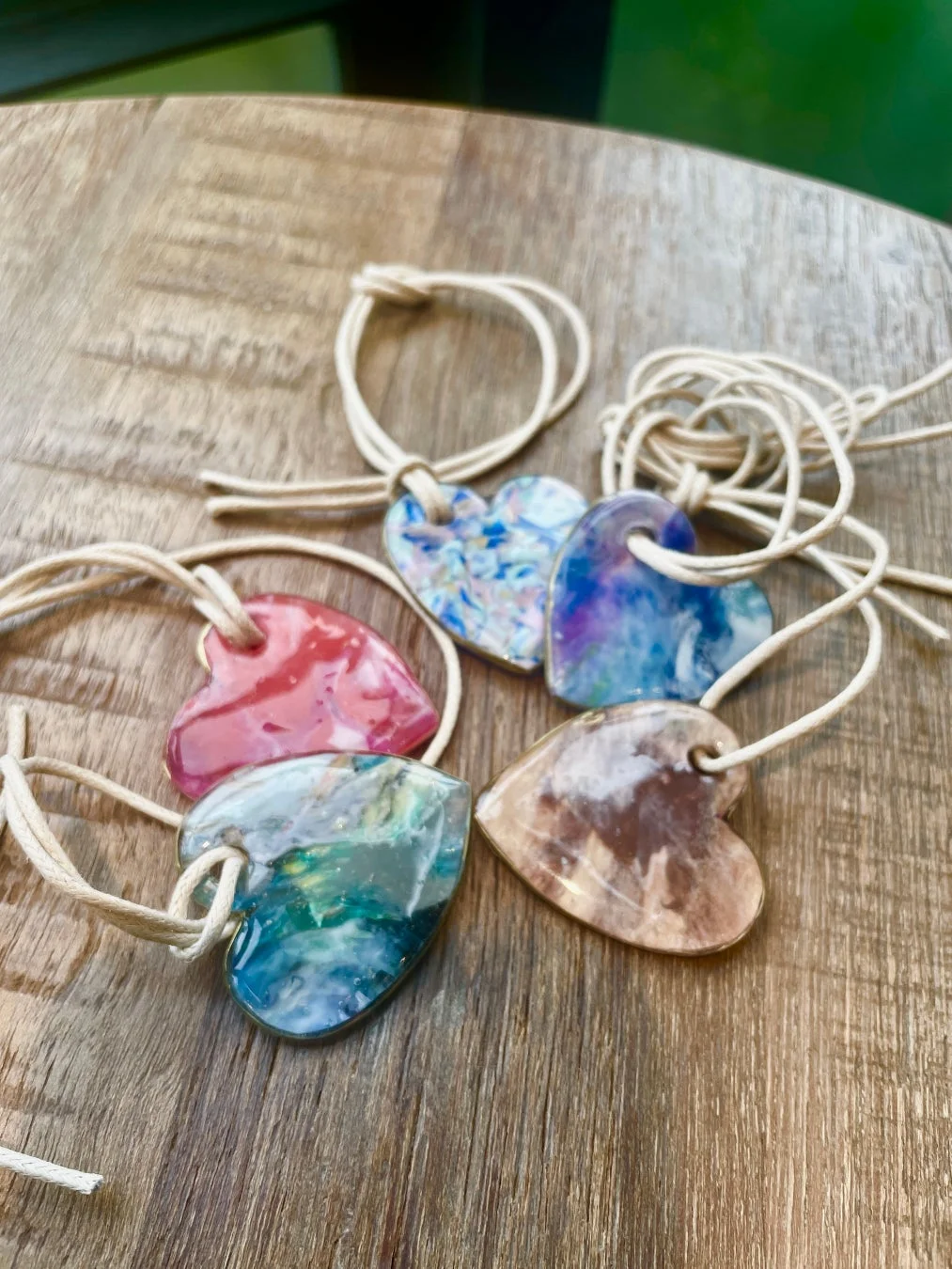 木製のテーブルに、紐が通されたカラフルなハート型のペンダントが複数置かれています。それぞれが異なる色合いの美しいマーブル模様をしており、手作りのアクセサリーのようです。