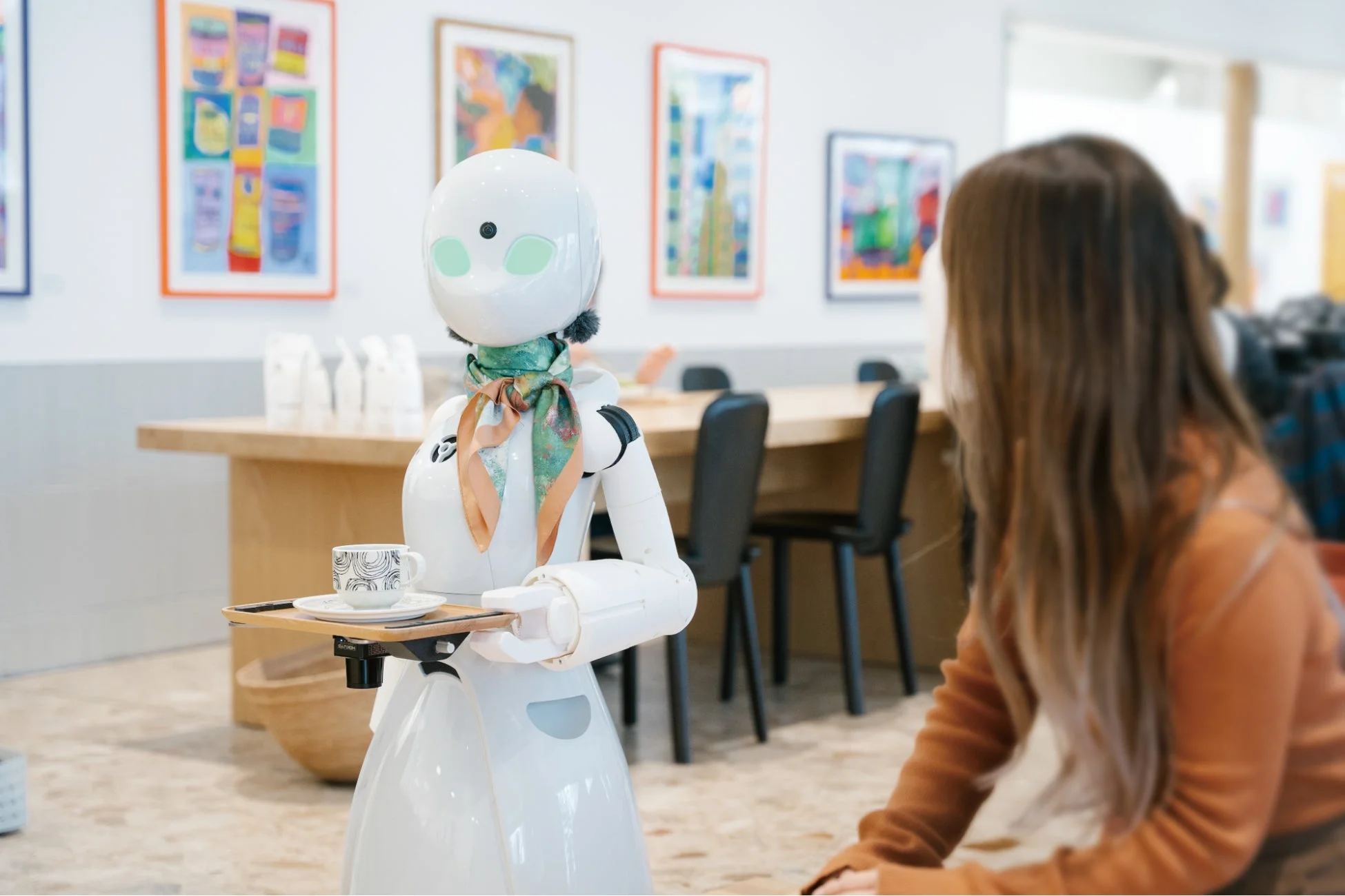 スカーフを巻いた白いロボットが、コーヒーカップが乗ったトレイを運び、座っている客の方を向いている様子。