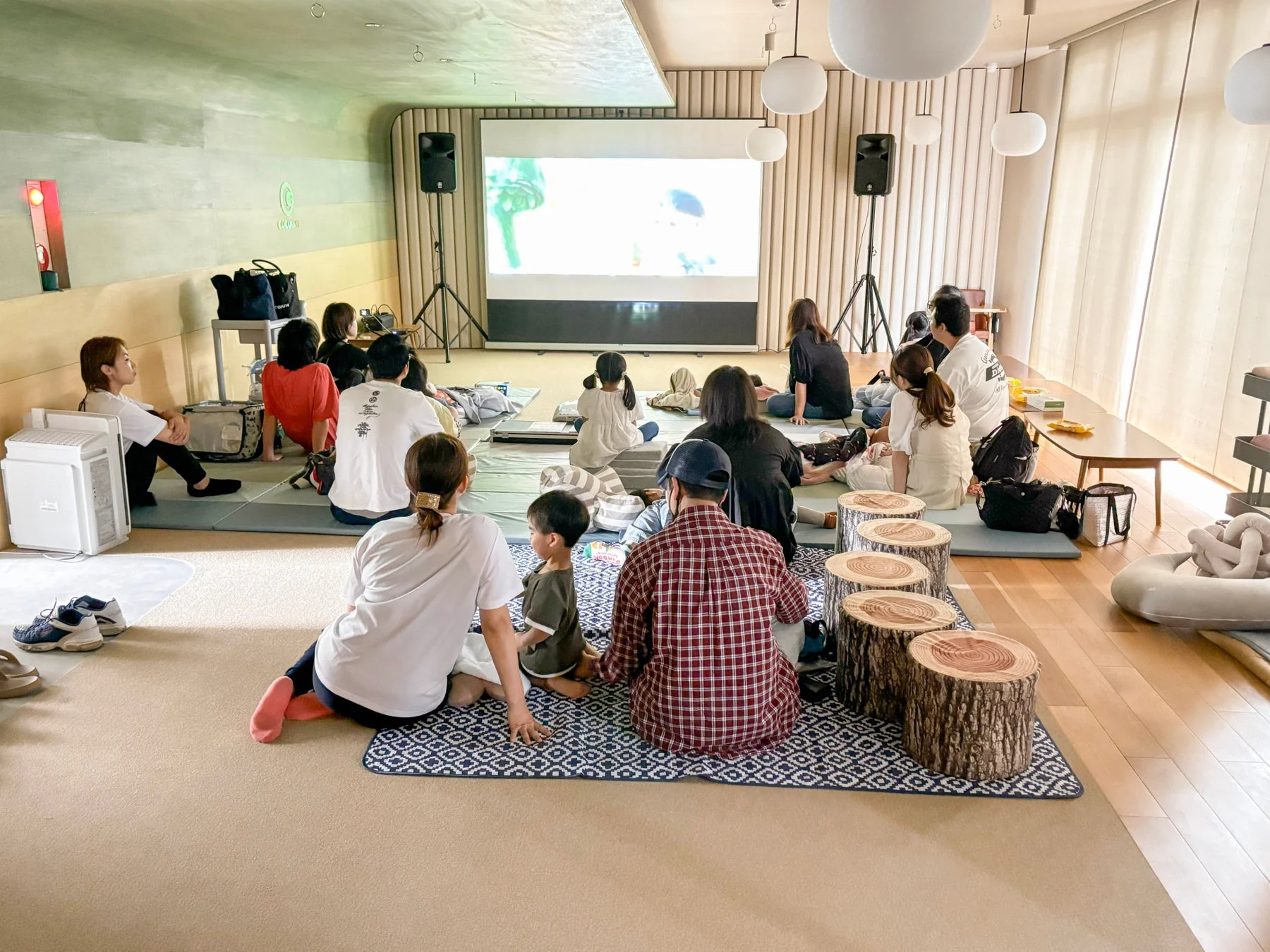 多くの人々が屋内の広々とした空間で、床に座って大きなスクリーンに映し出された映像を鑑賞しています。子供連れの家族が多く、リラックスした雰囲気の中でイベントを楽しんでいる様子がうかがえます。