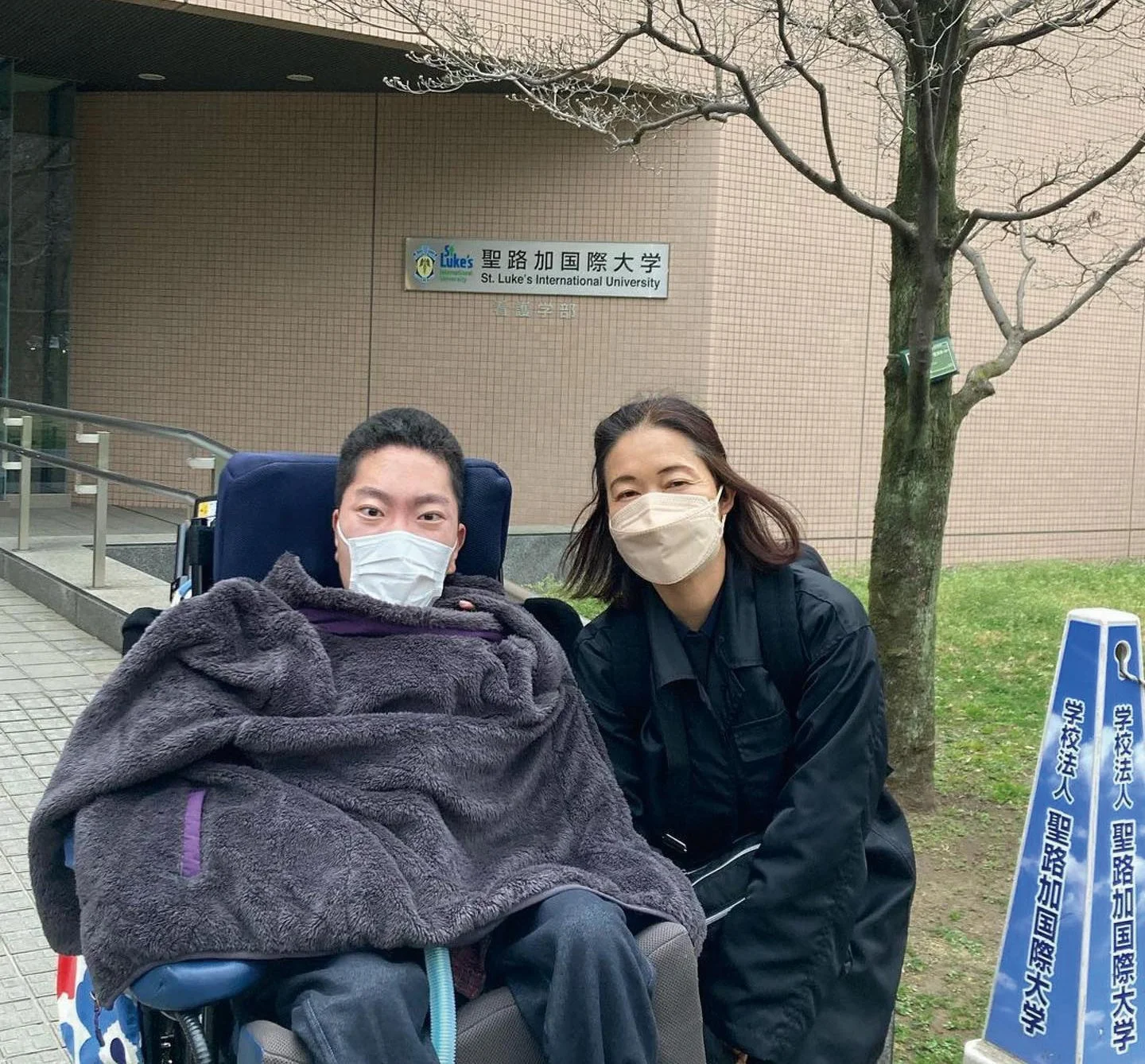 聖路加国際大学の前で、車椅子に乗った男性と女性がマスク姿で笑顔を見せている