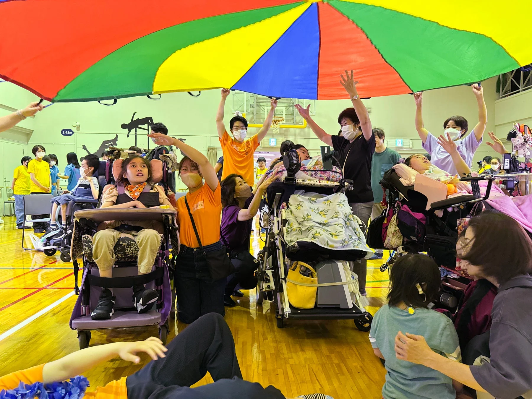 体育館で、大人と子供たちがカラフルなパラシュートを使って遊ぶレクリエーション活動の様子