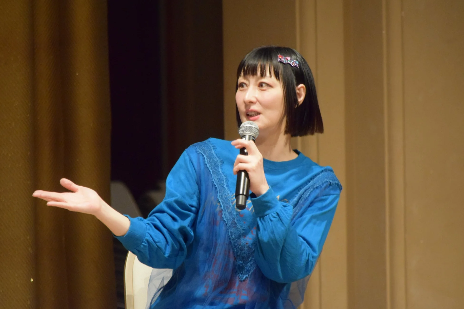 青い服を着た女性がマイクを手に持ち、ステージ上で話している様子。ショートヘアで、左手でジェスチャーを交えながら講演やプレゼンテーションを行っている場面を捉えた一枚です。