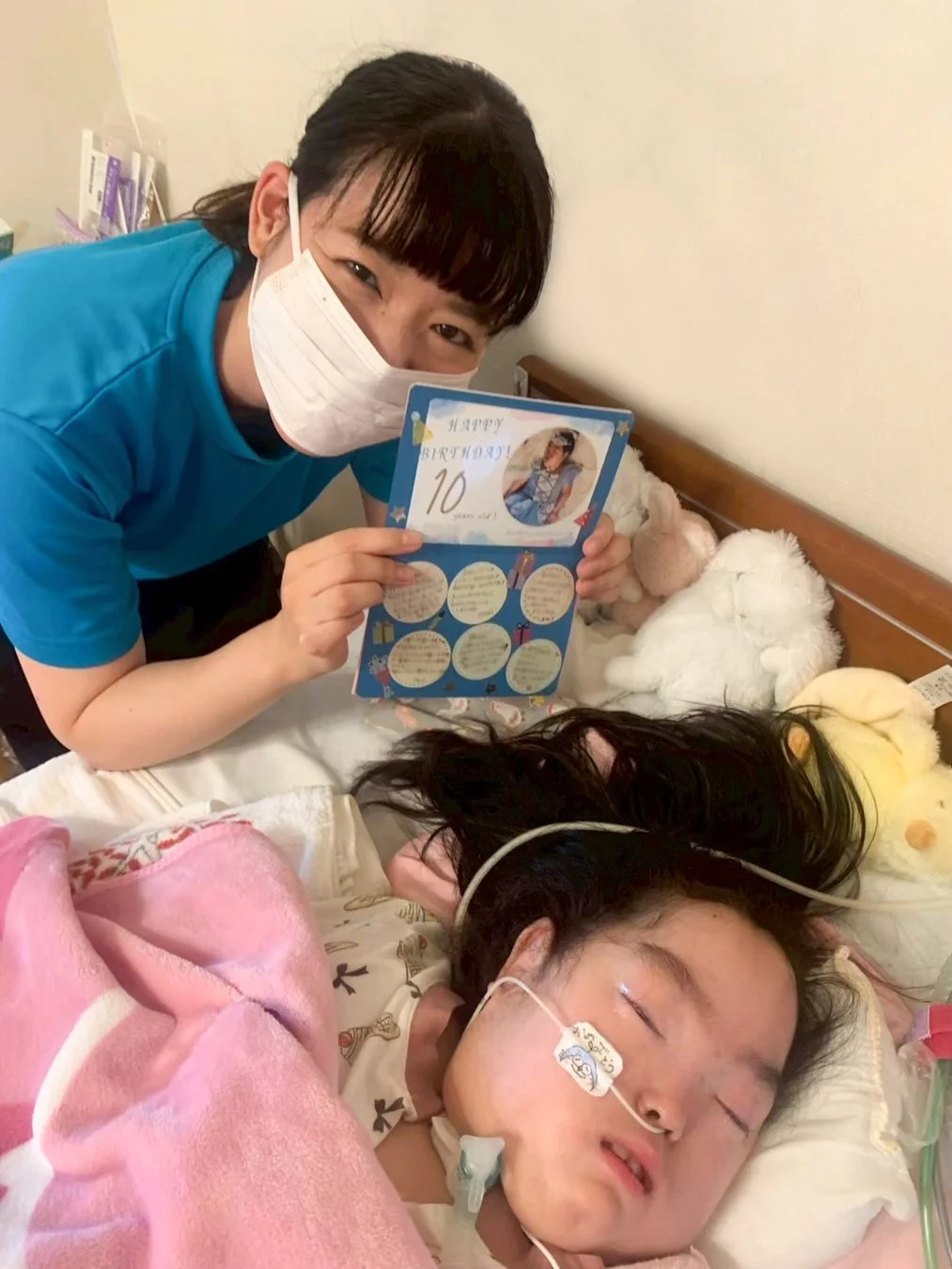マスクをした女性がベッドに横たわる子供のそばで「HAPPY BIRTHDAY! 10 years old!」と書かれたバースデーカードを見せている