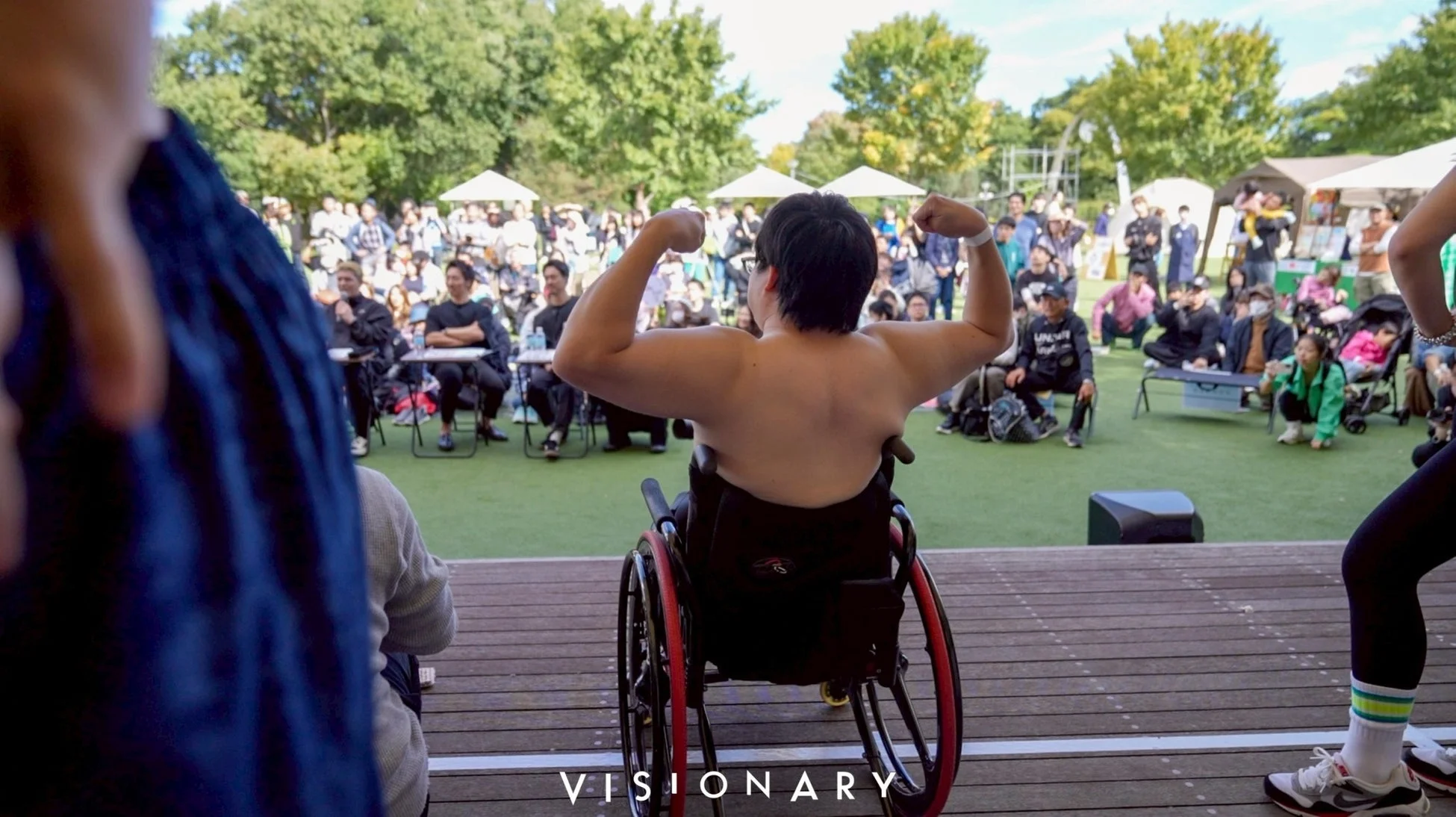 屋外イベントのステージ上で、車椅子に乗った人が背中を見せて力こぶを作っている様子
