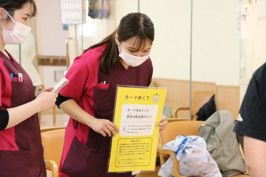 マスクを着用した2人の女性スタッフが室内にいる。1人が「カードめくり」という活動の説明が書かれた黄色の看板を持っている。保護者向けのメッセージも含まれており、施設内でのアクティビティや説明会の様子。
