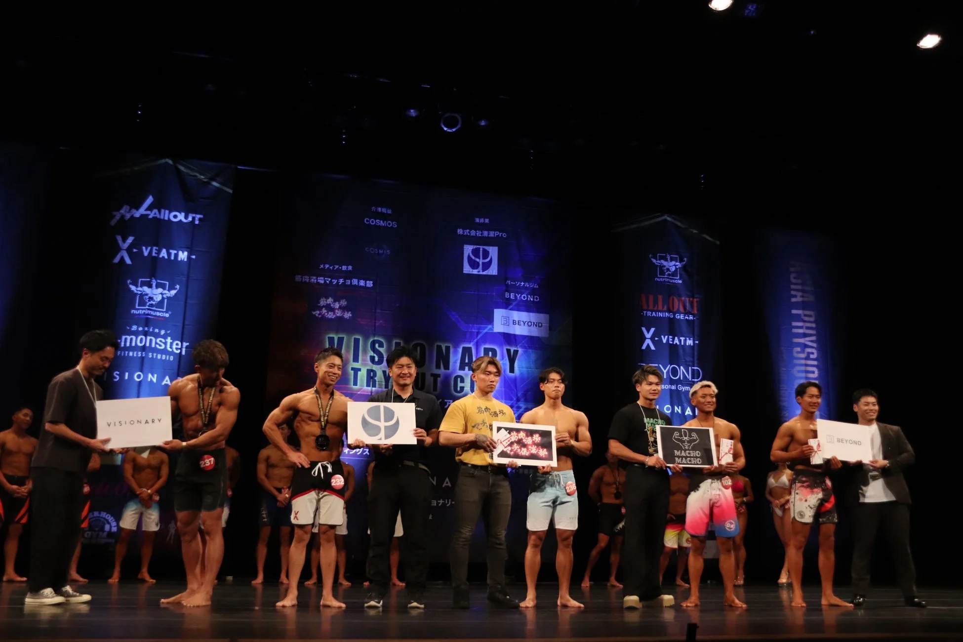 フィットネスコンテストの表彰式で、複数の男性ボディビルダーがステージ上でメダルやトロフィーを手にしています