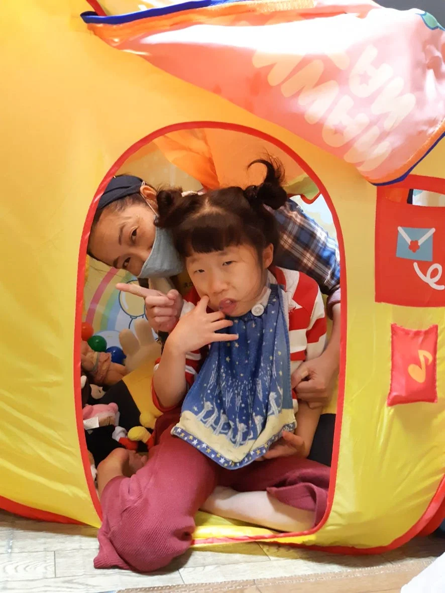 黄色いプレイテントの中で、おかっぱの子供と大人が楽しそうに遊んでいる