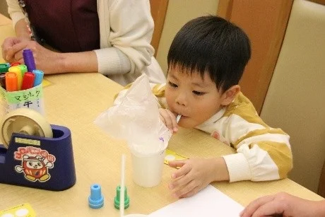 幼い男の子がテーブルで何かに集中して取り組んでいます。プラスチックの袋やコップ、ストローを使い、周りにはマーカーやテープディスペンサーなどの画材が見えます。