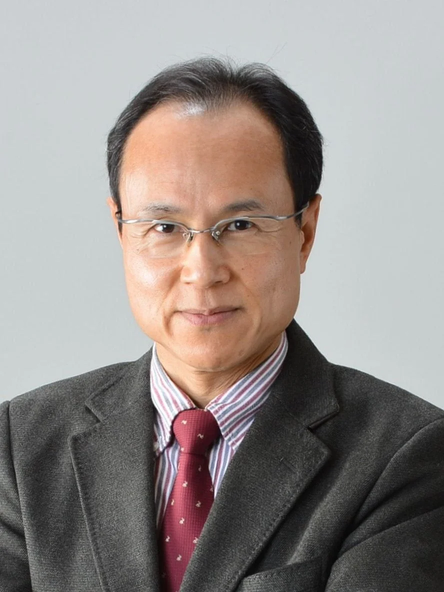 眼鏡をかけたアジア系の男性が、スーツジャケットとストライプシャツに赤いネクタイを着用し、カメラをまっすぐ見つめている上半身のポートレート写真です。