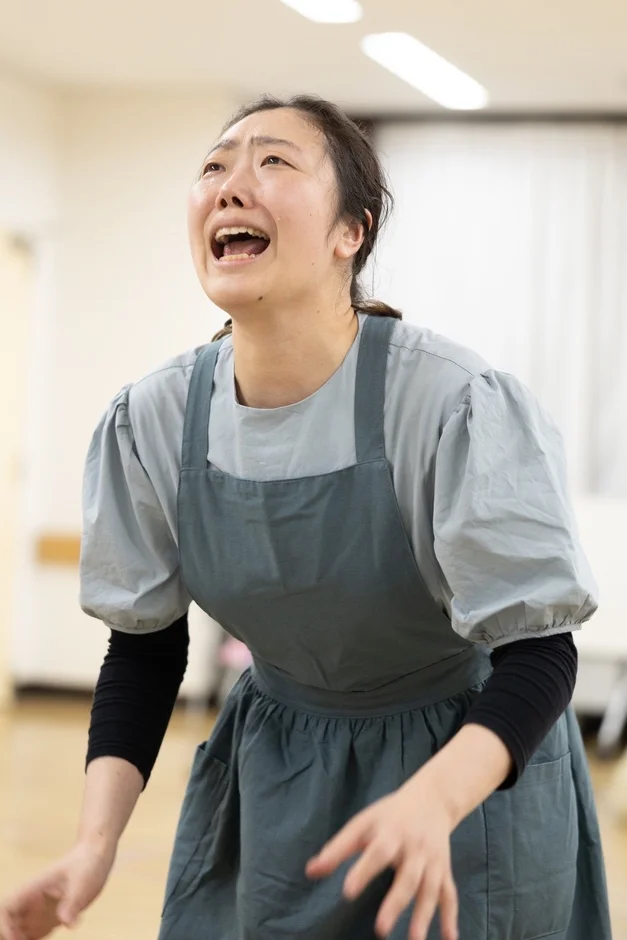 エプロンを着用した若い女性が、顔を上に向けて激しく泣き叫んでいる様子。涙が頬を伝い、口を大きく開けており、強い悲しみや絶望といった感情が表現されているドラマチックな瞬間を捉えた画像です。