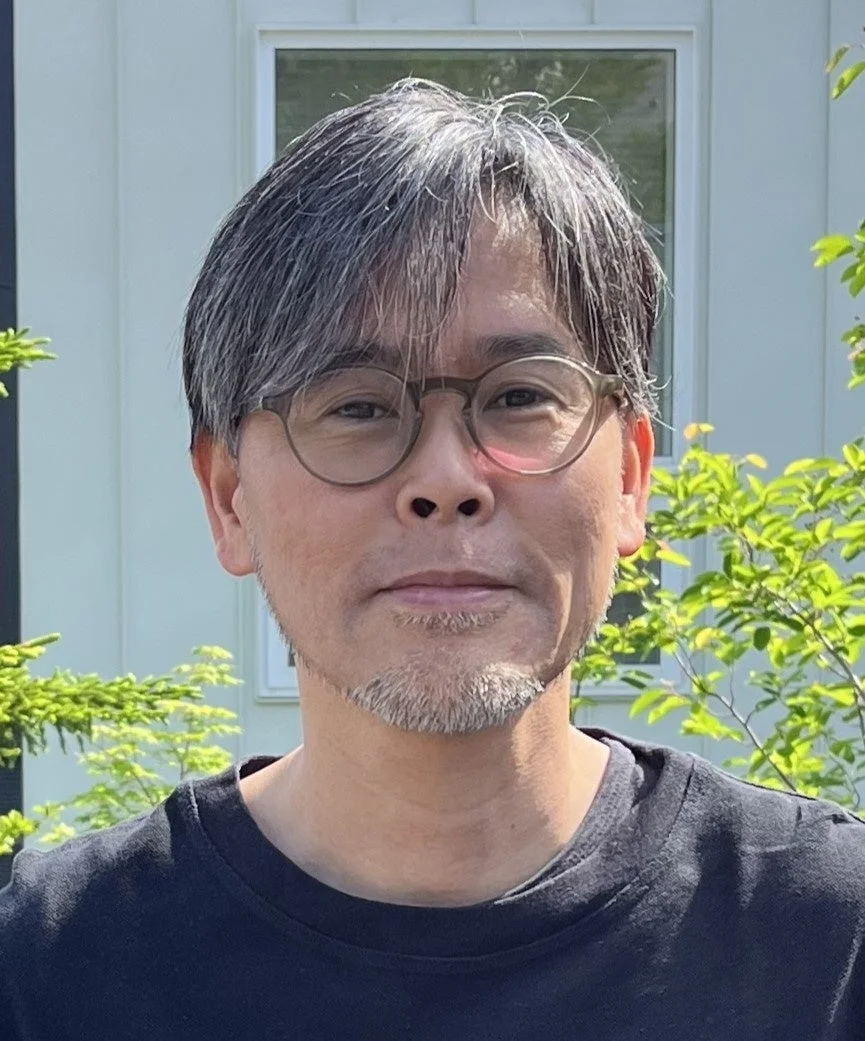 眼鏡をかけた中年のアジア人男性が、屋外で白い家と緑を背景に立っています。カジュアルな黒いTシャツを着用し、白髪混じりの髪と顎髭が特徴的なポートレートです。