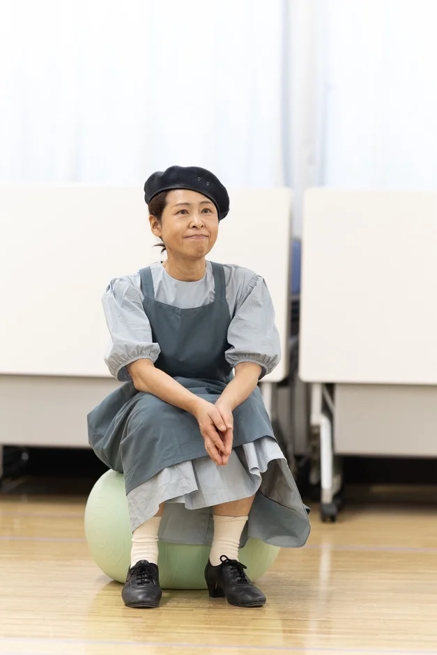 ベレー帽とエプロンドレスを着た女性が、ライトグリーンのバランスボールに座っています。穏やかな表情で前方を見ており、屋内でのリラックスした様子がうかがえます。