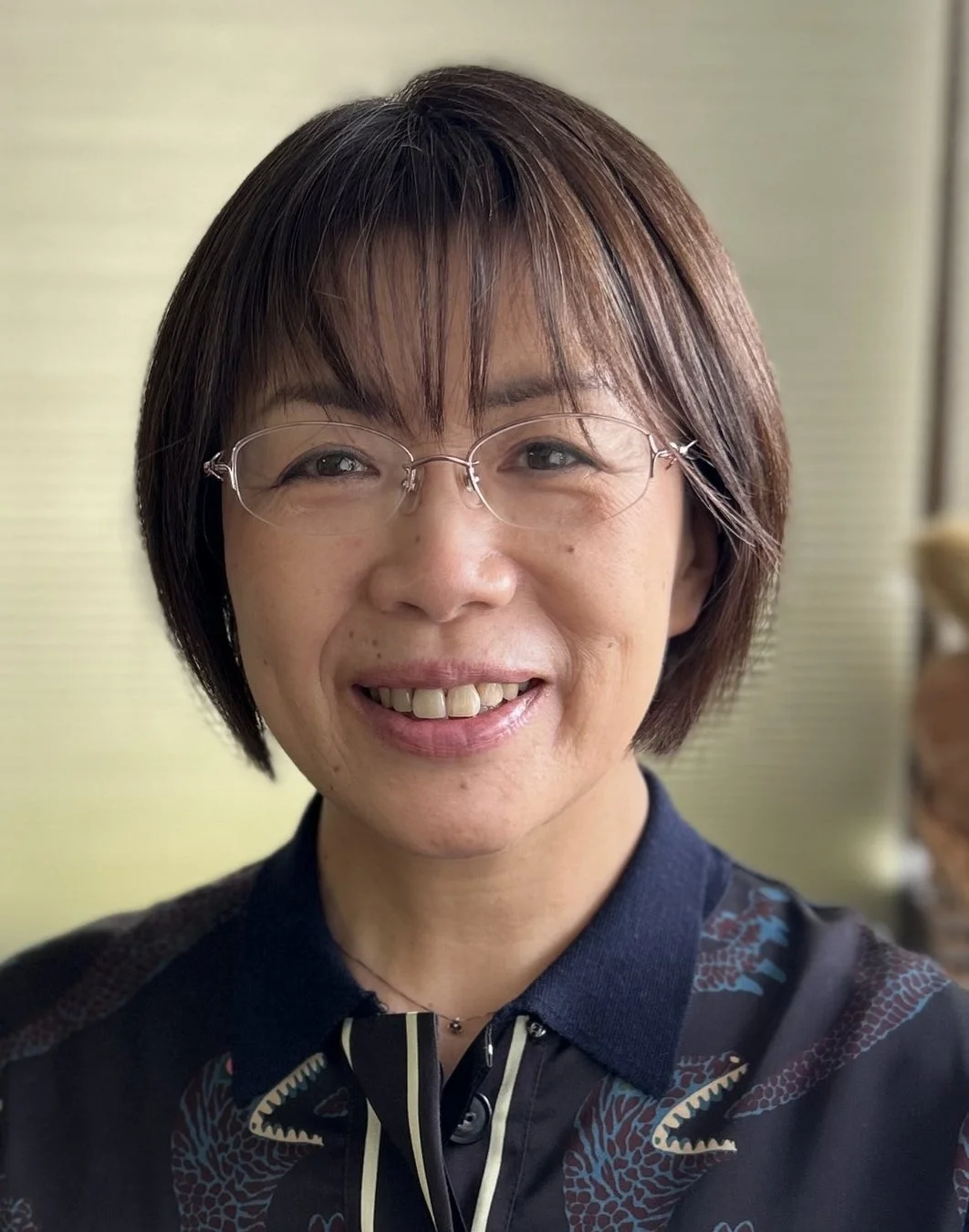 眼鏡をかけたショートヘアの女性が、カメラに向かって優しく微笑んでいます。ダークカラーの柄入りシャツを着用し、親しみやすい表情です。