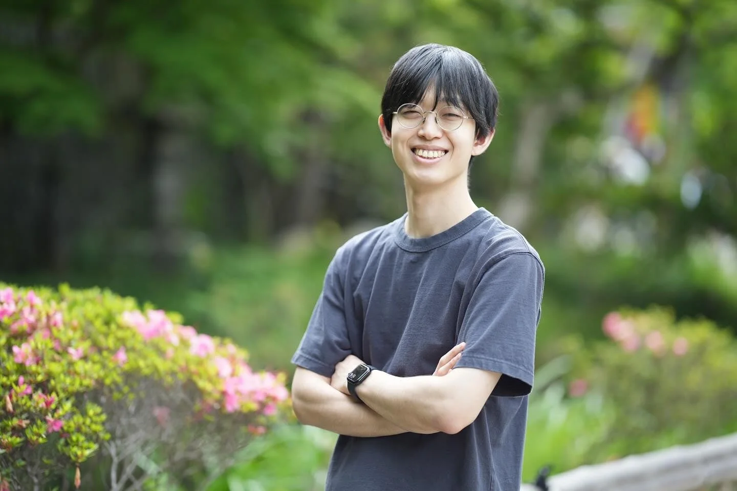 緑豊かな背景とピンクの花を背に、眼鏡をかけた若いアジア人男性が明るく微笑んでいます。彼は濃い色のTシャツを着て腕を組んでおり、爽やかな印象です。