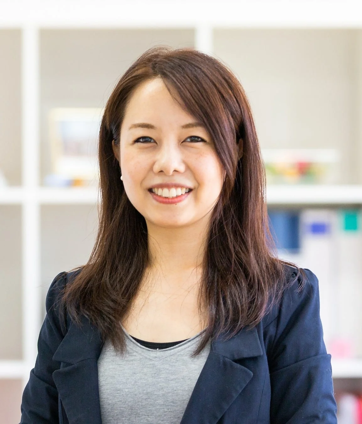 笑顔でカメラを見つめるアジア人女性のポートレート。紺色のブレザーとグレーのTシャツを着用し、背景はぼかされたオフィスのような空間です。