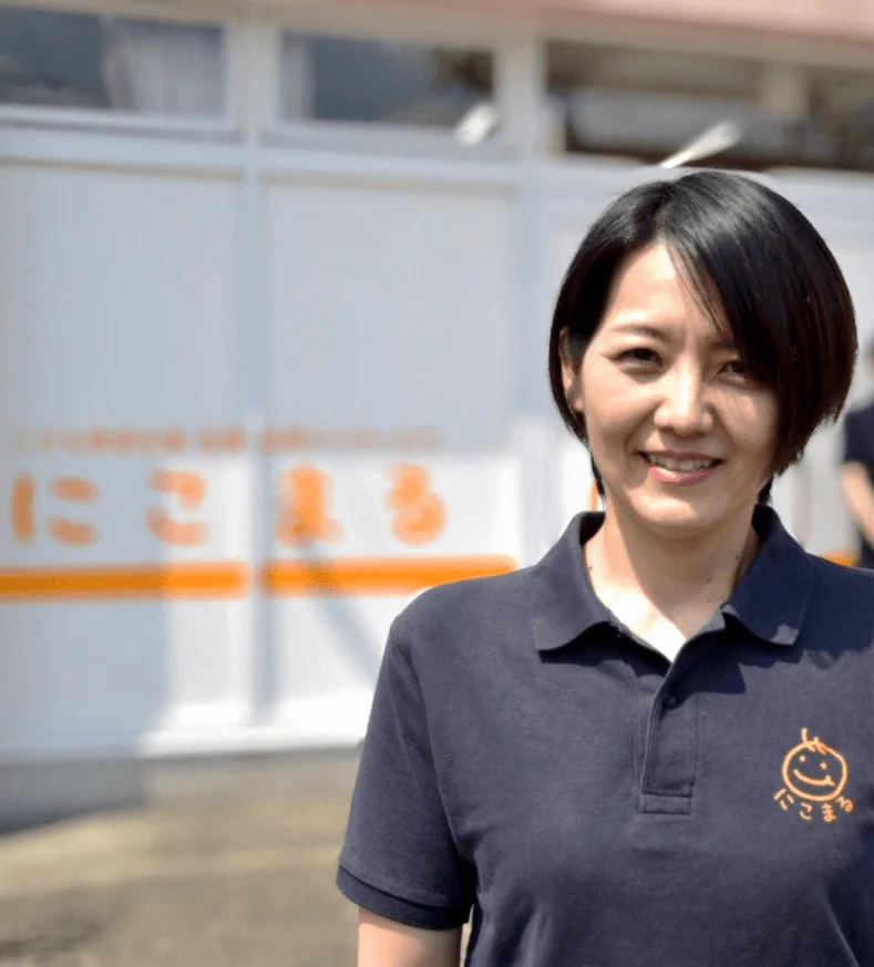 笑顔の日本人女性が「にこまる」のロゴ入り紺色ポロシャツを着て屋外に立っています