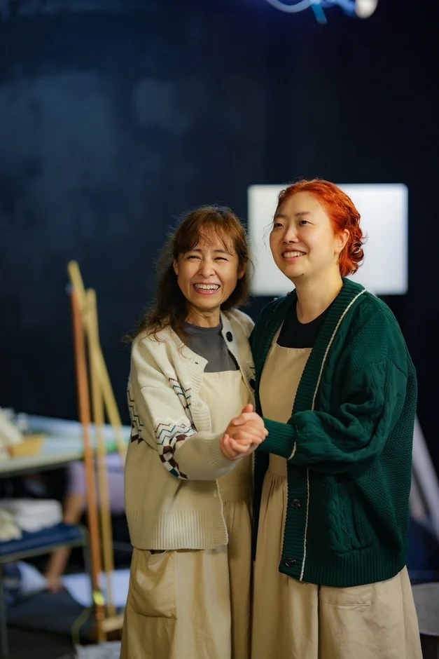 笑顔の2人の女性が手を取り合って立っており、楽しそうな雰囲気です。ベージュのエプロンとカーディガンを着用し、親密な絆を感じさせます。背景は暗く、スタジオのような場所で撮影されたようです。