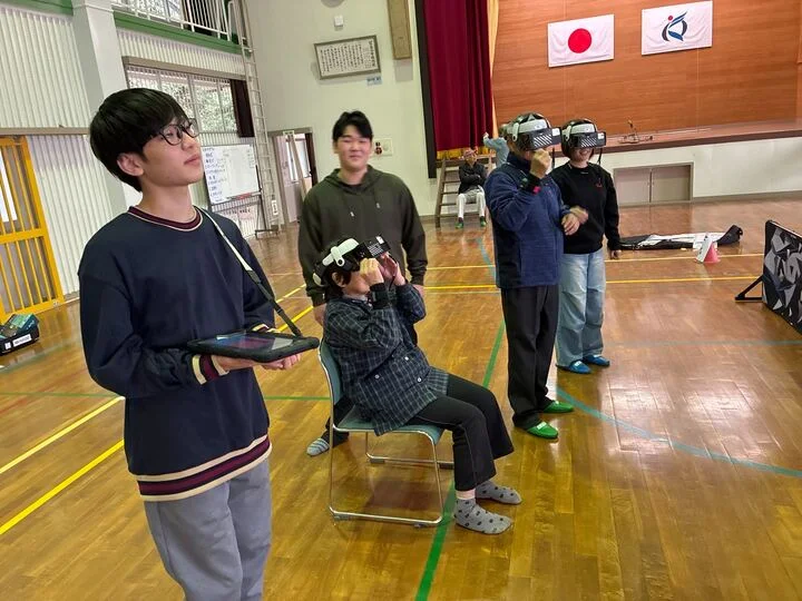 体育館のような場所で、数人がVRヘッドセットを装着してバーチャルリアリティを体験している様子です。