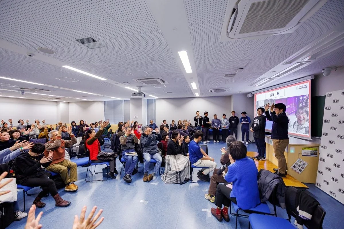 ボランティアサミットらしきイベントで、多くの参加者が手を挙げて活発に交流している様子