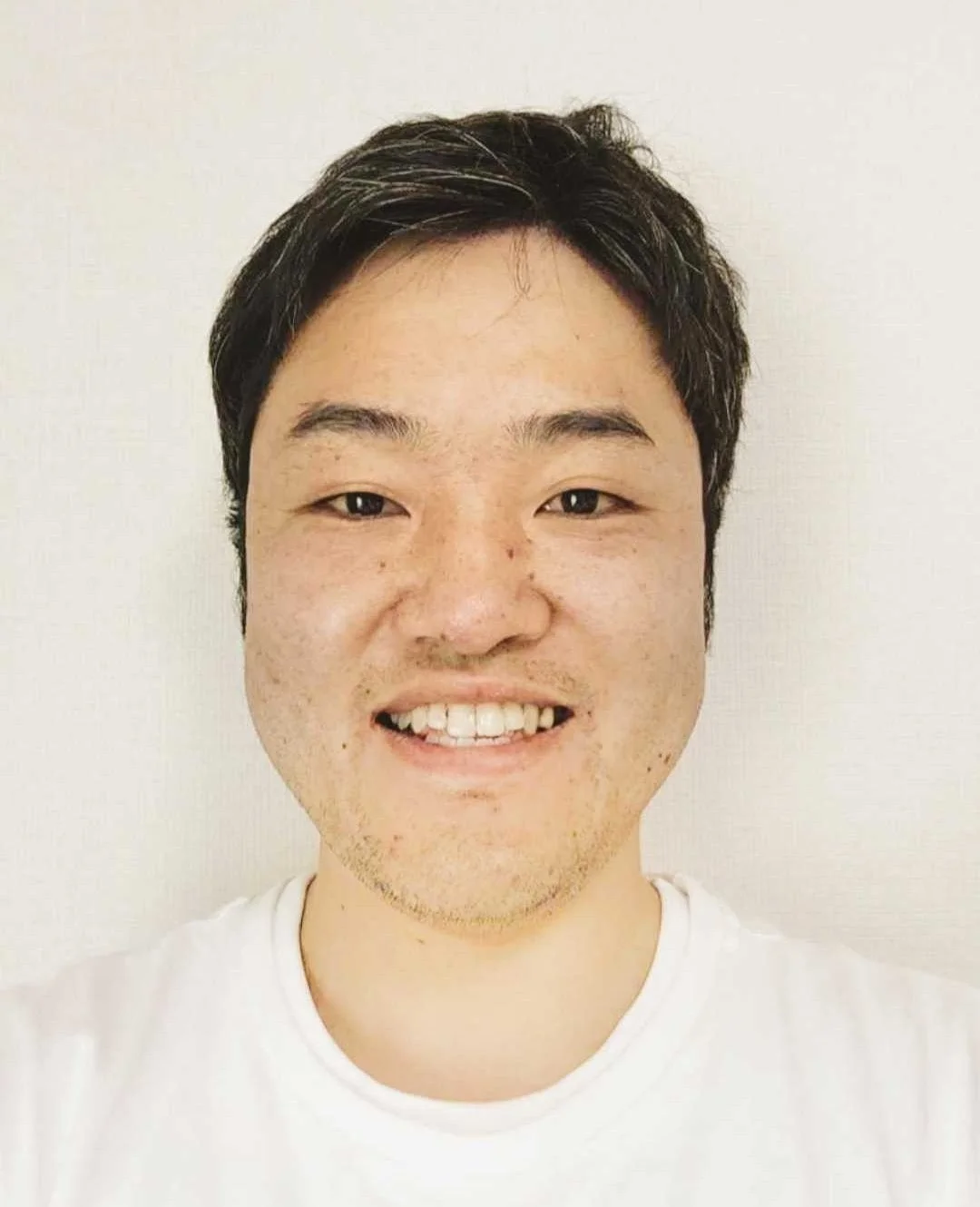 白いTシャツを着たアジア人男性の顔のアップです。彼はカメラに向かって穏やかに微笑んでおり、親しみやすい表情をしています。