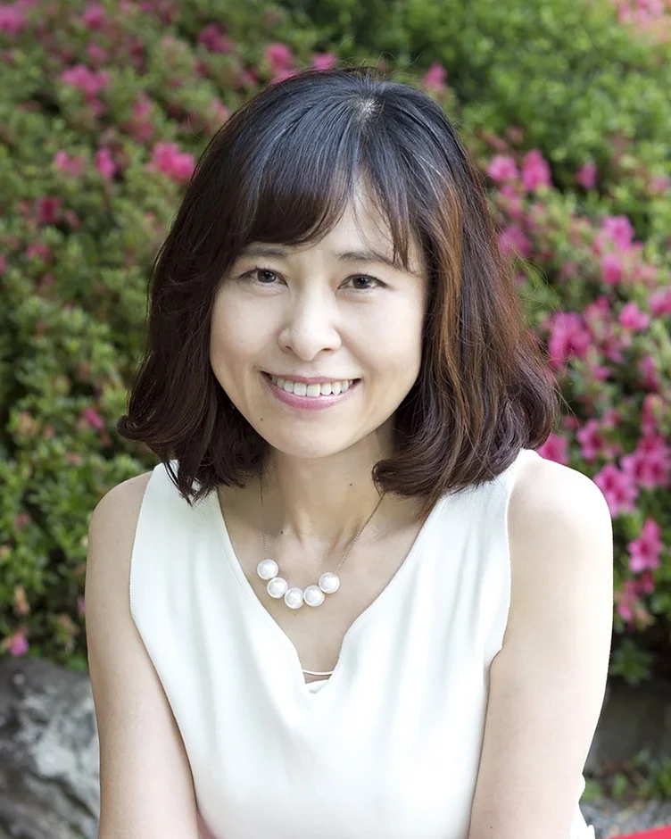 笑顔でカメラを見つめる日本人女性のポートレートです。彼女はショートヘアで白いノースリーブのトップスを着ており、真珠のようなネックレスをしています。背景にはぼかされた緑の葉とピンクの花が見えます。