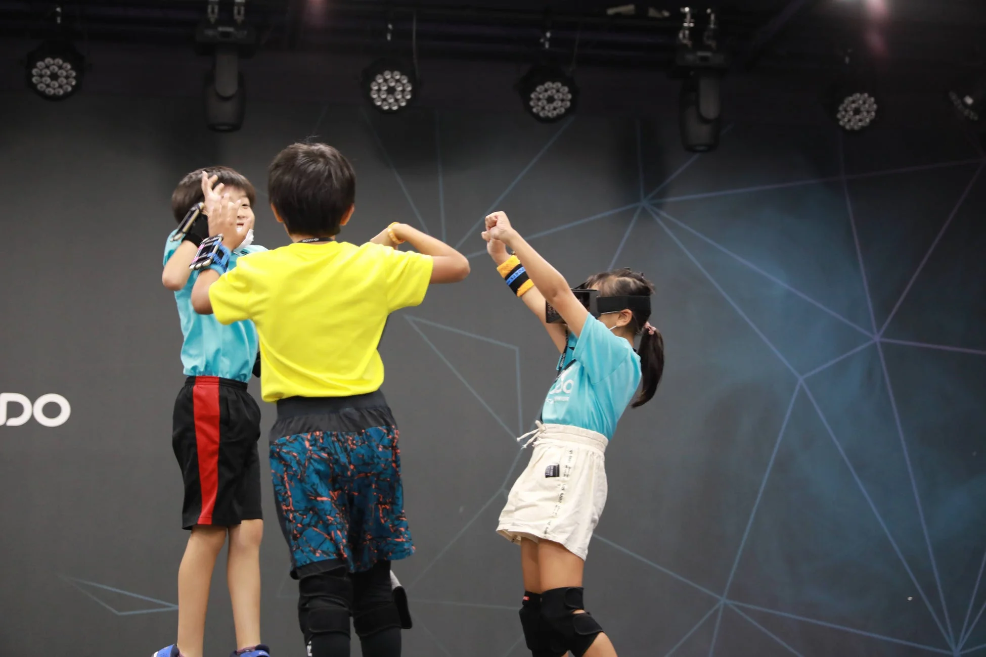 子供たちがVRヘッドセットやアームバンドを装着し、ステージ上で未来的なテクノロジーを使ったインタラクティブなゲームや体験を楽しんでいます。