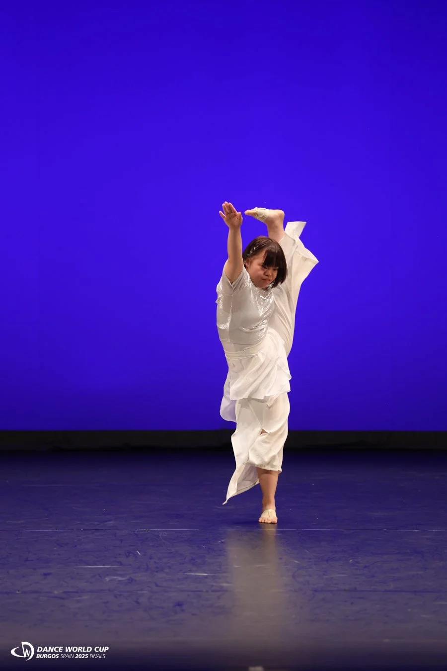 青い背景のステージで、白い衣装を着たダンサーが片足を高く上げた柔軟なポーズを披露している