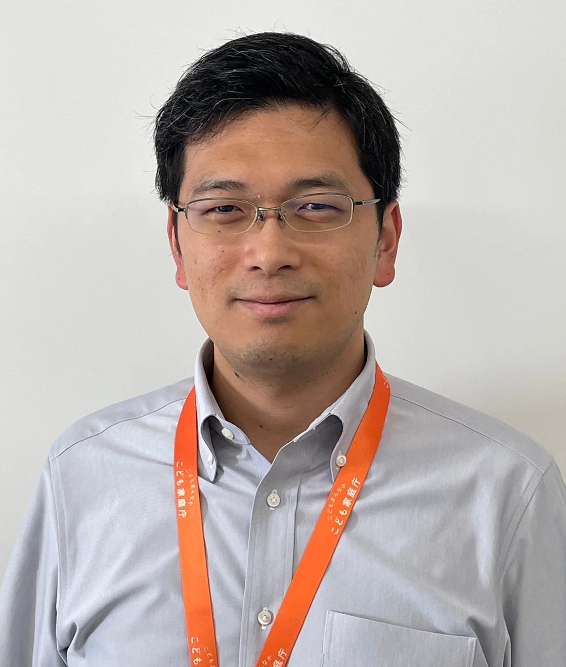 眼鏡をかけたアジア人男性のポートレートです。水色のシャツを着ており、首には「こども家庭庁」と書かれたオレンジ色のネックストラップを下げています。