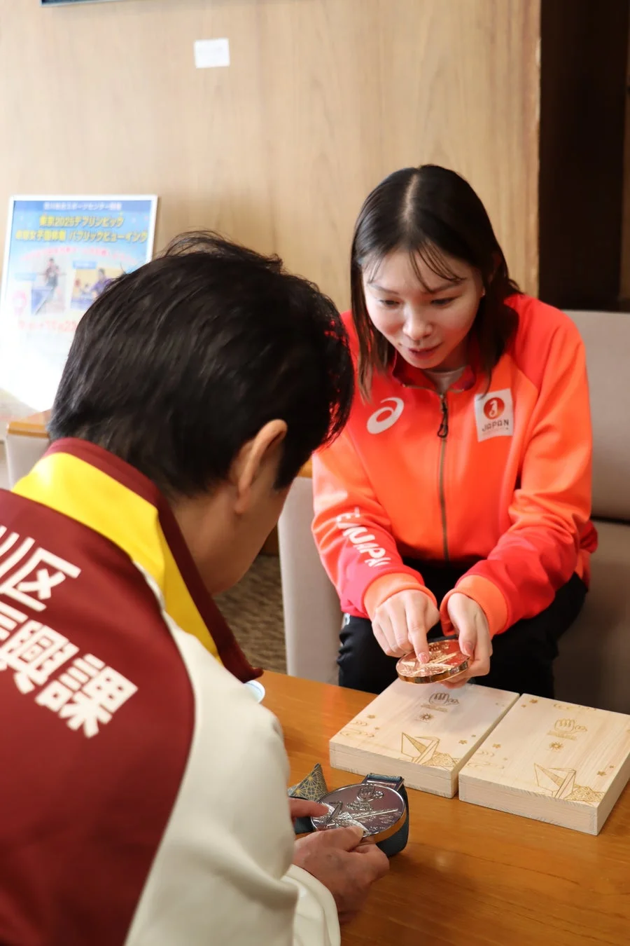 オレンジ色のJAPANジャージを着た女性が、男性にメダルを見せています