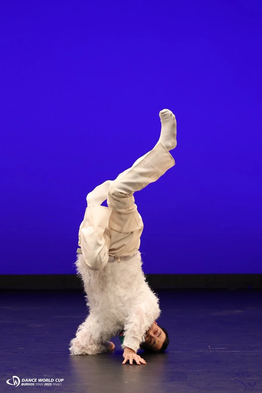 青い背景の舞台で、白いふわふわの衣装を着たパフォーマーがヘッドスタンドをしている