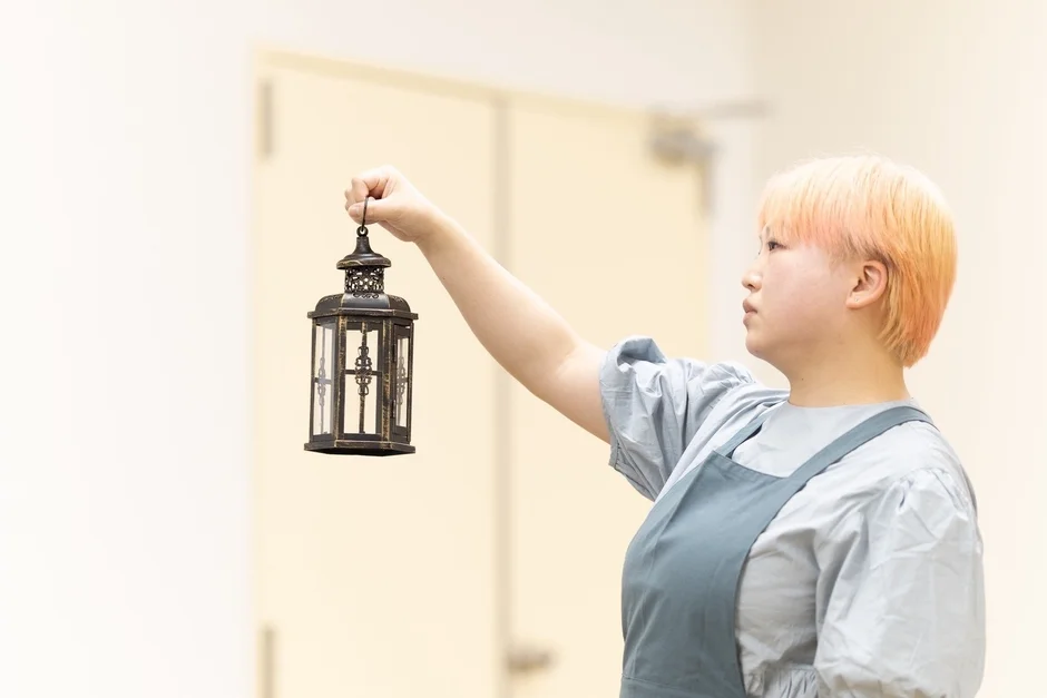 オレンジ色のショートヘアの女性がエプロンを着用し、アンティーク調のランタンを高く掲げています。明るい室内で横顔を見せており、何かを見つめているような雰囲気です。