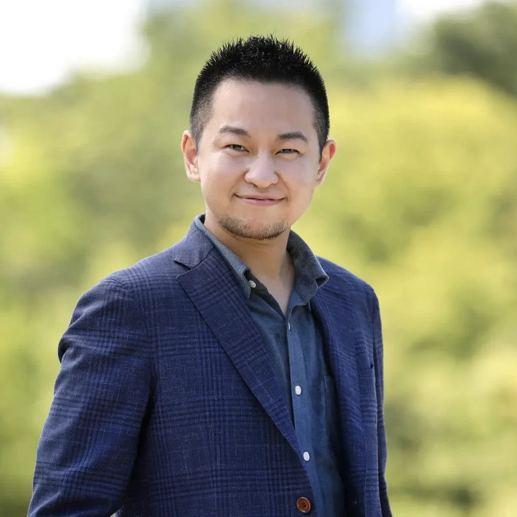 青いチェックのブレザーとデニムシャツを着たアジア人男性のポートレート。屋外の緑を背景に、カメラに向かって笑顔を見せています。ビジネスとカジュアルを融合したスタイルです。