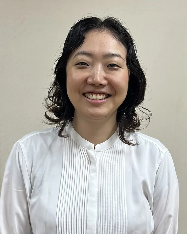 白いシャツを着たアジア人女性が、カメラに向かって笑顔で写っているポートレート写真です。背景はシンプルな無地で、清潔感のある印象を与えます。