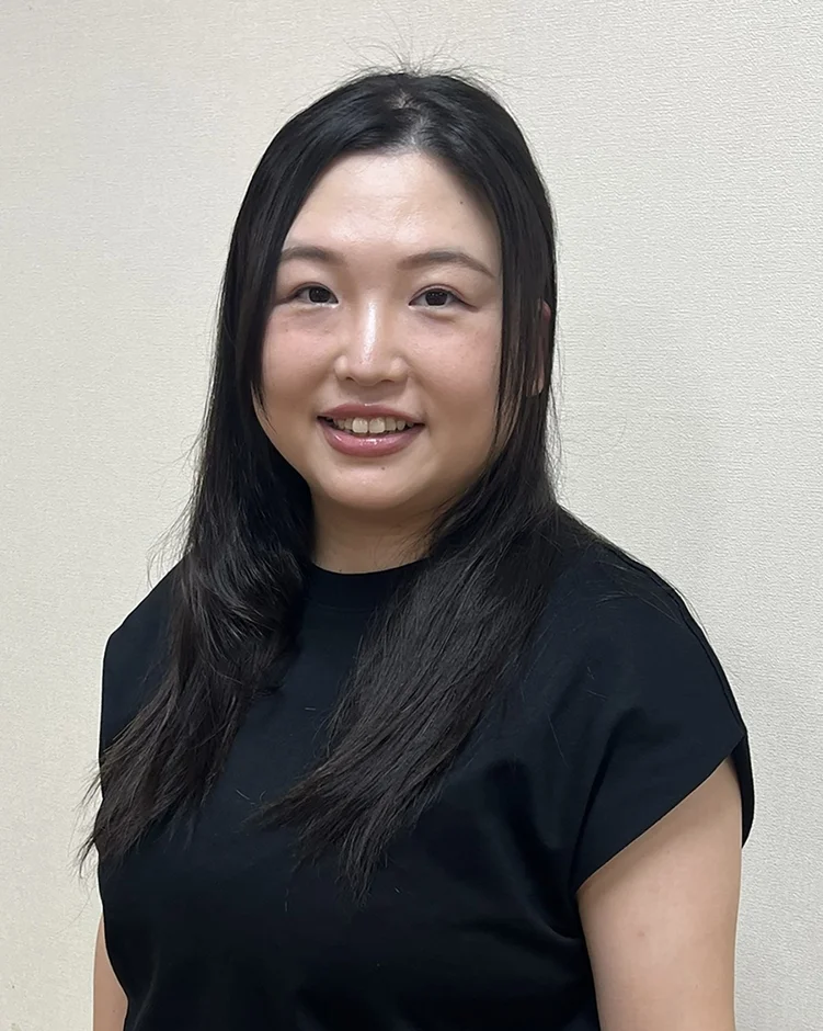 黒いTシャツを着たアジア人女性のポートレート。長い黒髪で、カメラに向かって優しく微笑んでいます。背景はシンプルな無地です。