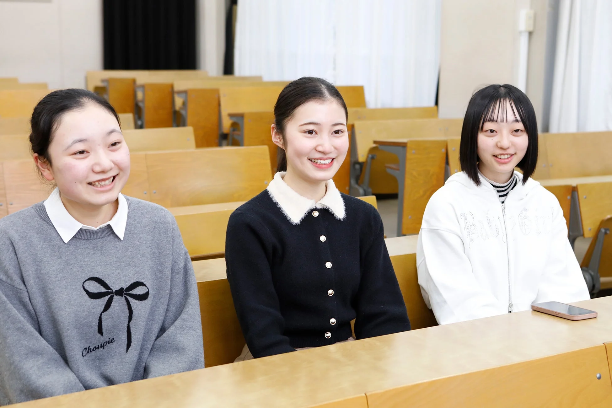 講義室のような場所で、3人の若い女性が笑顔で座っています。彼女たちはカジュアルな服装をしており、机の上にはスマートフォンが置かれています。