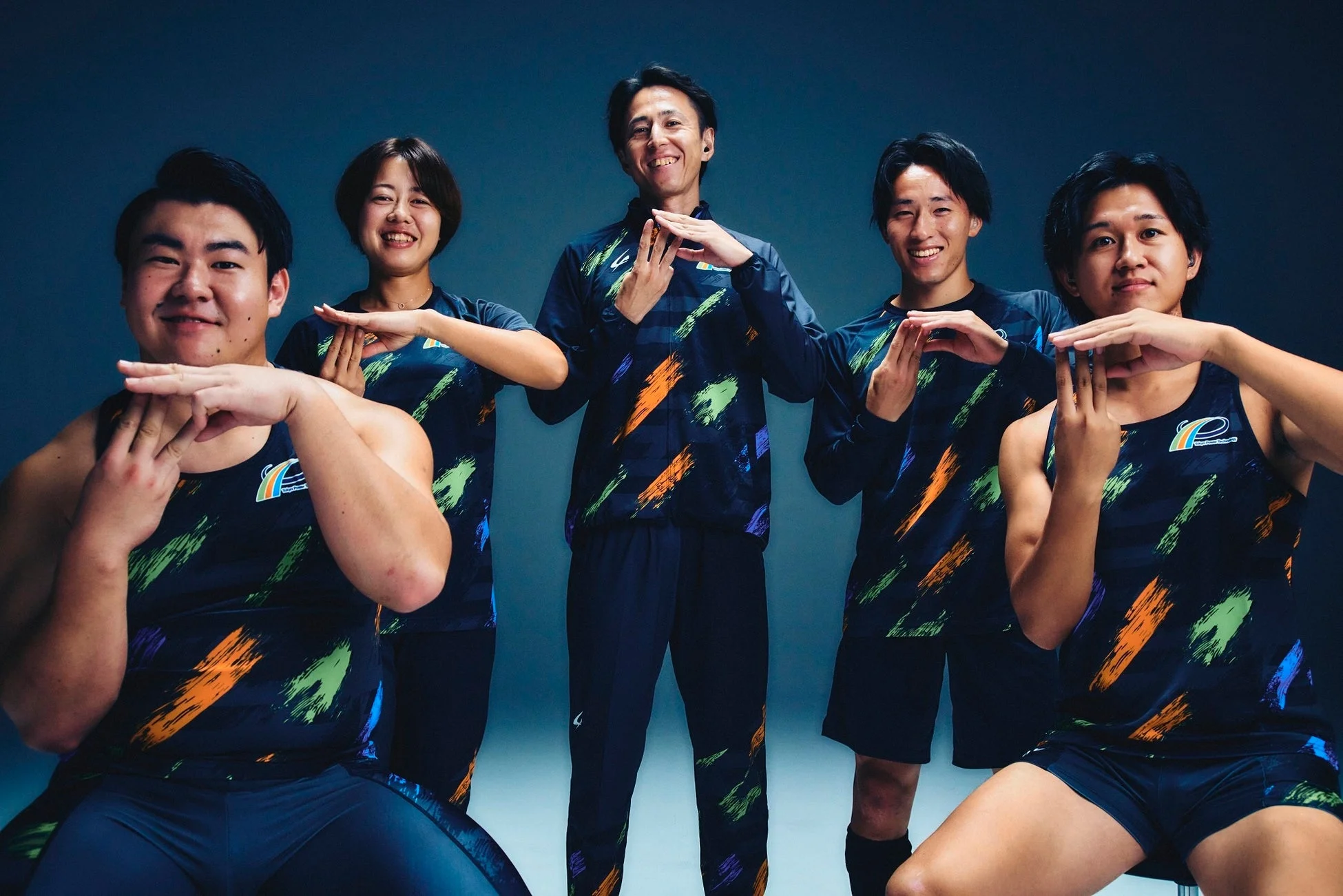 カラフルなブラシストローク柄のユニフォームを着用した男女5人のアスリートが、笑顔で同じ手を使ったポーズをとっています。