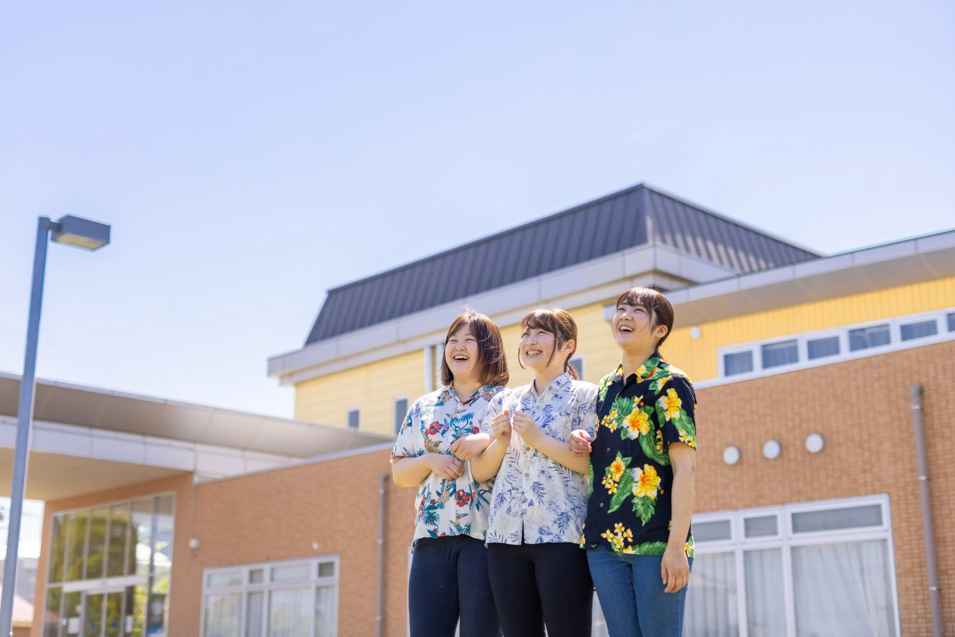晴れた屋外で、アロハシャツを着た3人の女性が上を向いて楽しそうに笑っている