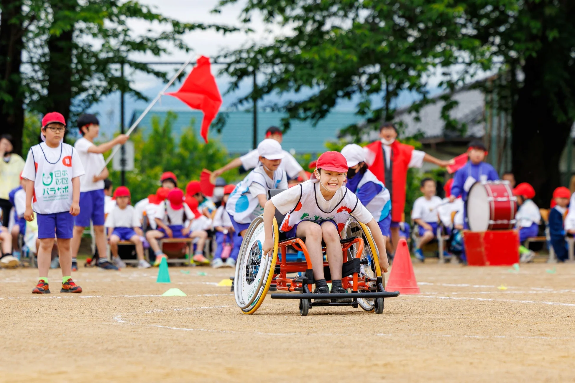 運動会のようなイベントで、笑顔の子供が競技用車椅子に乗って楽しそうにレースに参加しています。他の子供たちや大人が見守る中、パラスポーツの楽しさを伝える活気ある瞬間が捉えられています。