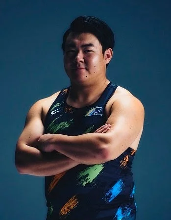 青い背景の前で、カラフルなタンクトップを着用し、腕を組んで立つ筋肉質な男性のポートレート。